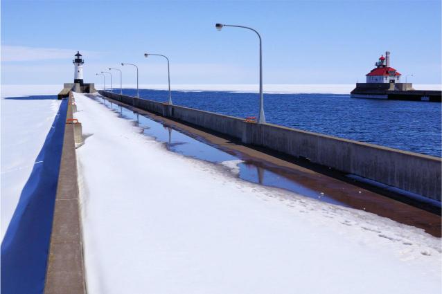 A photograph featuring a snowy pier extending out into a frozen lake, with a lighthouse on the left and a ship in the distance.