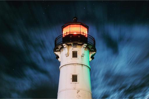 A lighthouse at night with clouds in the background, illuminated by a light on its top.