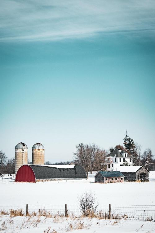 A snowy landscape featuring a red barn and farm buildings with silos in the background under a blue sky.