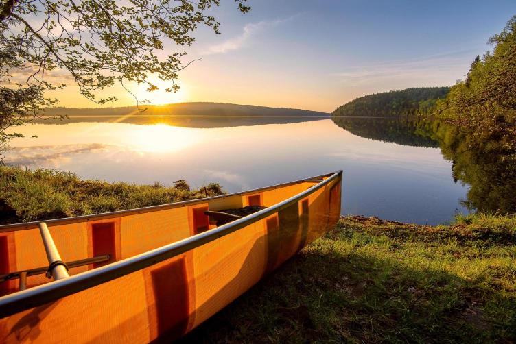 A canoe beached on the shore of a lake at sunset, with trees in the background.