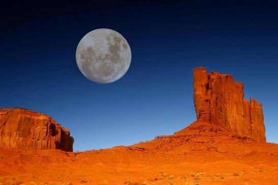 The image features a dramatic desert landscape with two prominent red buttes in the background against a clear blue sky. A large, bright full moon hangs in the sky, adding a serene and majestic quality to the scene.