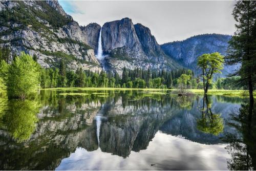 A scenic view of Yosemite National Park featuring a lake in the foreground with a reflection of the sky and mountains, a large waterfall in the background, and lush greenery around.