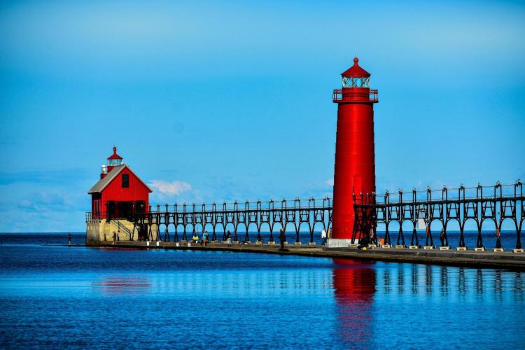 A red lighthouse with a covered walkway, located on a body of water under a clear blue sky.