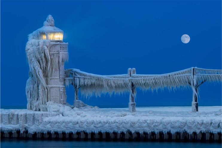 The image shows a frozen lighthouse and pier with ice covering everything, including the lamp of the lighthouse which is illuminated. The sky is clear and there is a full moon visible.