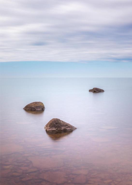 A tranquil lake scene with a few rocks partially submerged in the water, under a cloudy sky.