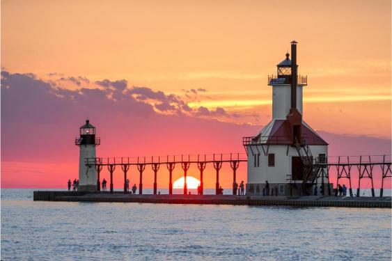 Two lighthouses on a pier at sunset with the sun low on the horizon, casting a pink and orange glow over the scene.