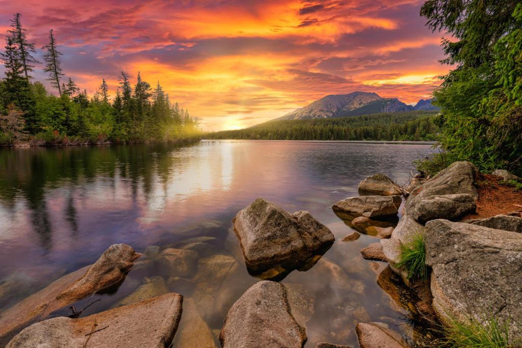 A scenic view of a lake at sunset with rocks in the foreground and trees lining the opposite shore.