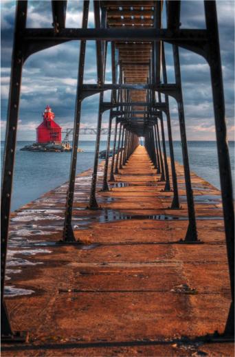 A wooden pier extending into water with a red lighthouse in the background and a partly cloudy sky above.