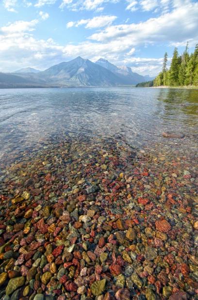 A clear lake with colorful rocks at the bottom, surrounded by trees and with mountains in the background under a blue sky with clouds.