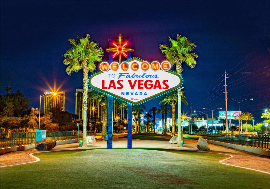 The iconic 'Welcome to Fabulous Las Vegas' sign at night, featuring a white background with blue and red text, illuminated by neon lights, with palm trees in the foreground.