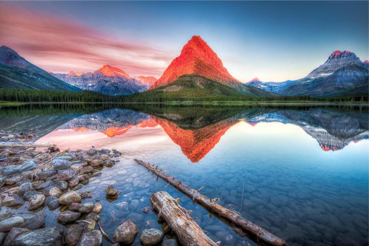 A scenic view of Swiftcurrent Lake in Glacier National Park at sunset, with a reflection of a red mountain peak on the water.
