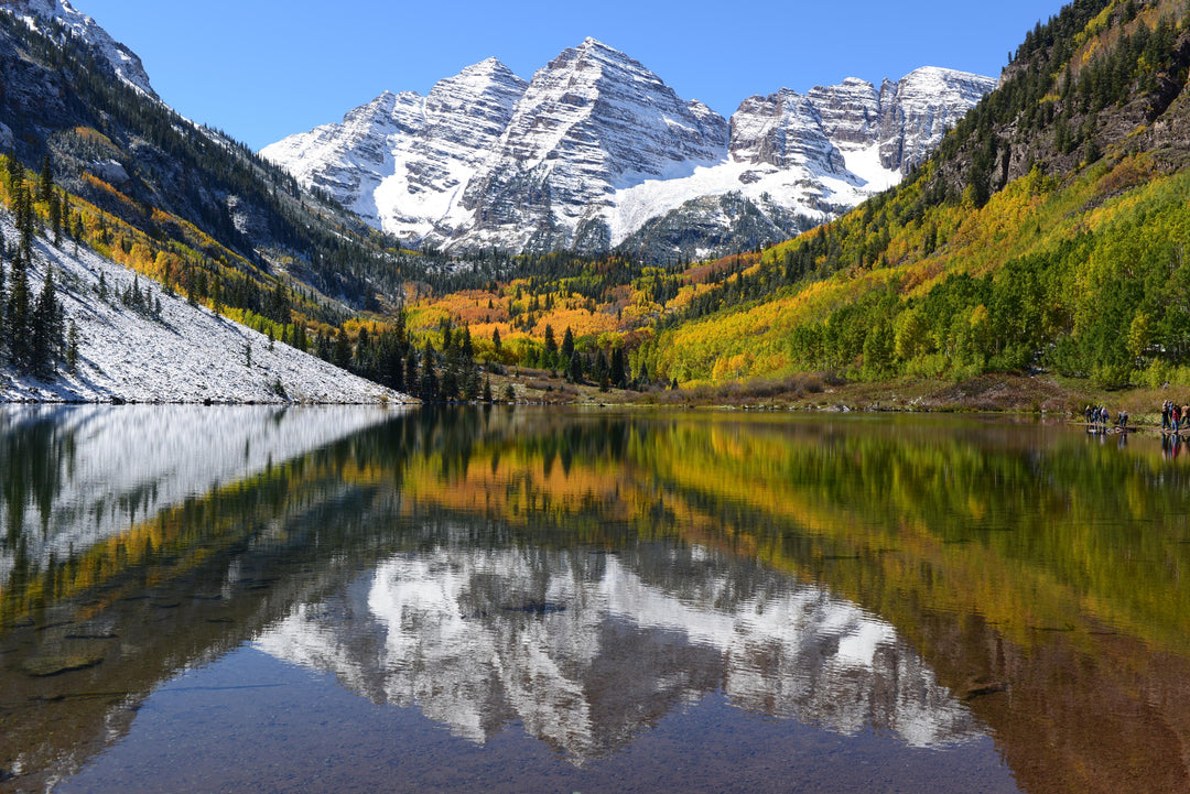 A scenic view of Maroon Bells in Aspen, Colorado, with snow-capped mountains reflecting in a still lake, surrounded by trees with autumn colors.