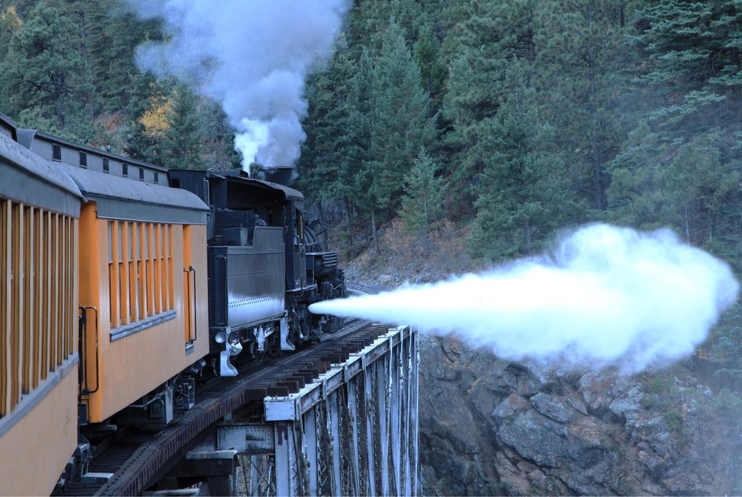 A steam engine train moving through a mountainous area, with billowing smoke from its chimney.