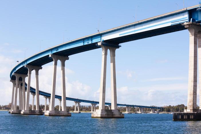 The image shows the San Diego Coronado Bridge with a clear blue sky in the background and water beneath it.