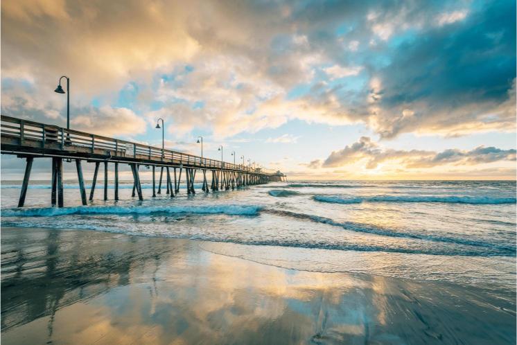 A pier extending over the ocean into the distance with a cloudy sky above and the ocean reflecting in the wet sand in the foreground.