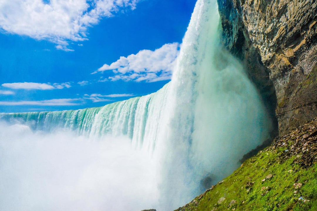 The image shows a partial view of Niagara Falls with a focus on the water's edge and the base of the falls, surrounded by some greenery on a bright, clear day.