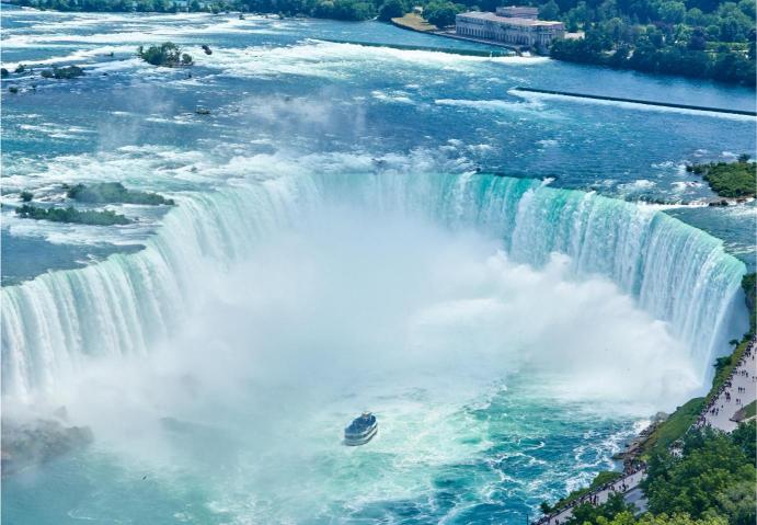 An aerial view of Niagara Falls with a small boat in the frame, showcasing the horseshoe shape of the falls and the mist created by the water.