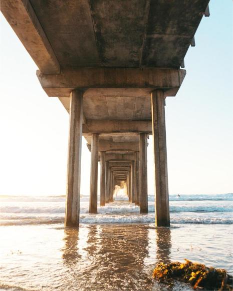 View looking down the length of a concrete pier at La Jolla, San Diego, with ocean waves and sunlight streaming through the support columns.