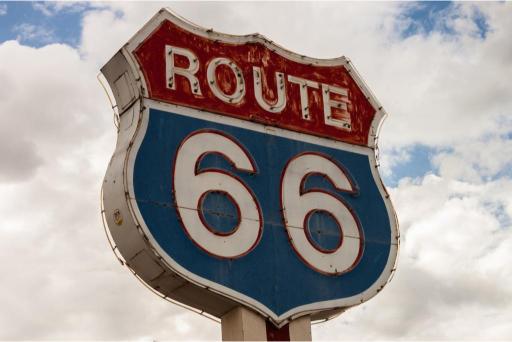 A signpost with 'ROUTE 66' written in white on a blue background, with a red 'ROUTE' banner on top against a cloudy sky.