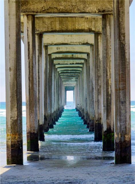 View under a large pier with several support columns, looking out towards the ocean.