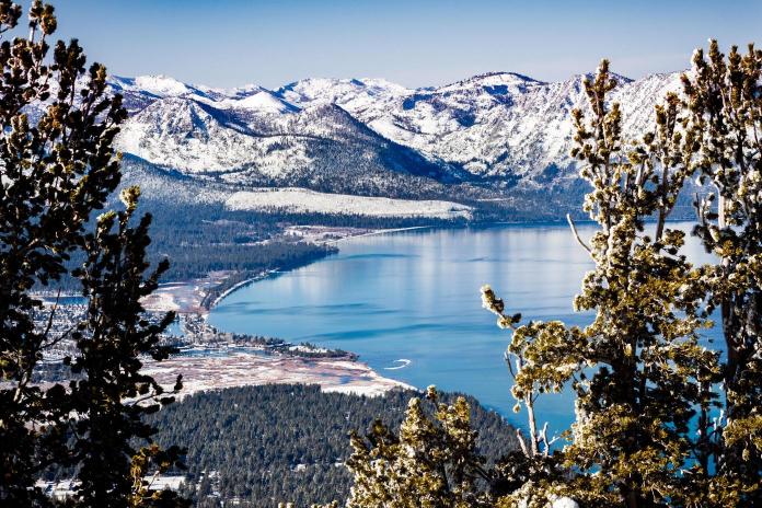 An aerial view of Lake Tahoe with snow-capped mountains in the background and evergreen trees in the foreground.