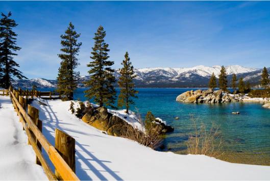 Winter landscape at Lake Tahoe with snow on the ground and a clear blue lake, surrounded by pine trees and mountains in the background.
