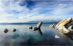 A clear blue lake with rocks partially submerged in the water, under a blue sky with few clouds.