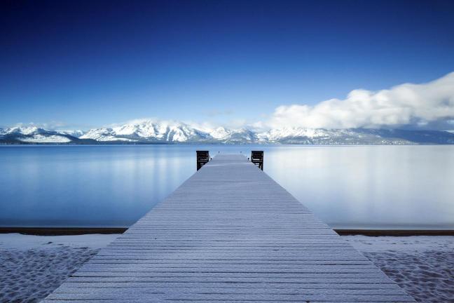 A snowy pier extending out over a lake with mountains in the background under a blue sky with clouds.