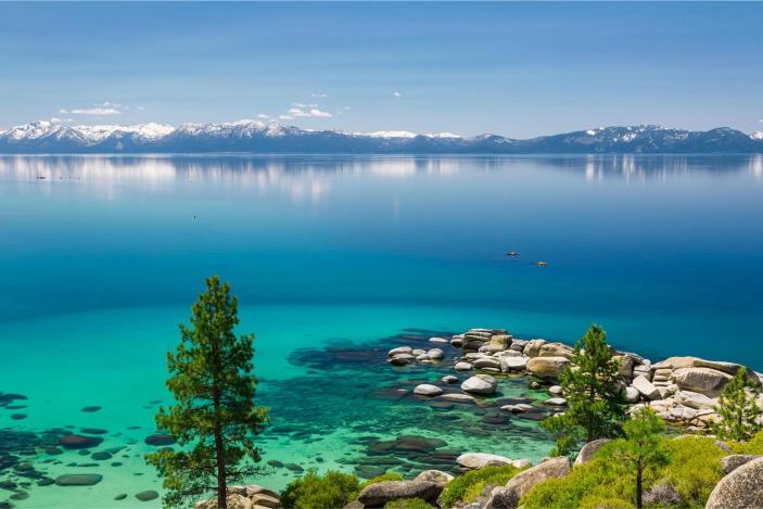 A clear blue lake with a mountain range in the background and a tree in the foreground