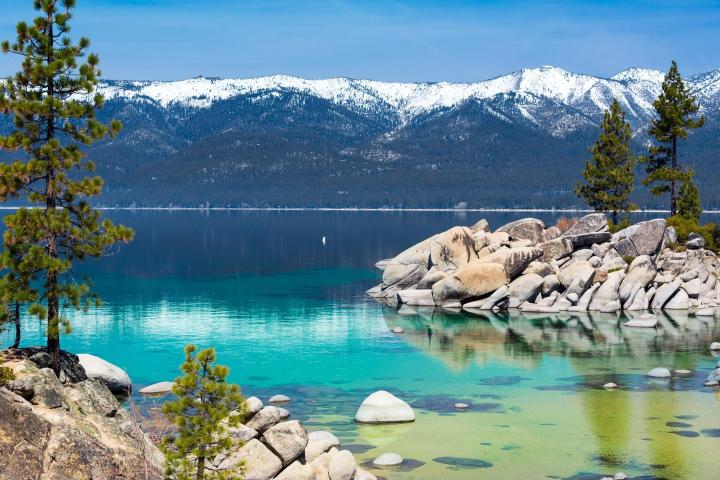 A clear blue lake with rocks and trees in the foreground, mountains with snow in the background, and a blue sky above.