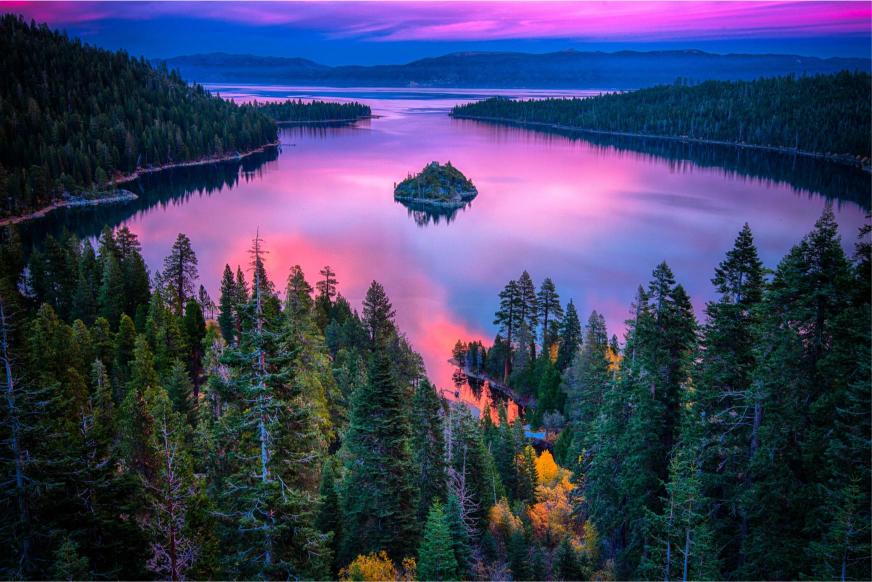 A scenic view of Lake Tahoe with the Sierra Nevada in the background, featuring a small island in the center and a pink and purple sky at sunset.