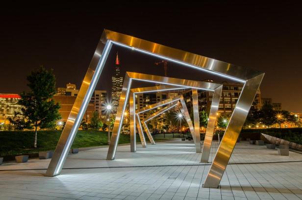 Metal sculpture consisting of multiple geometric shapes, likely a part of an art installation, with a cityscape in the background at night.
