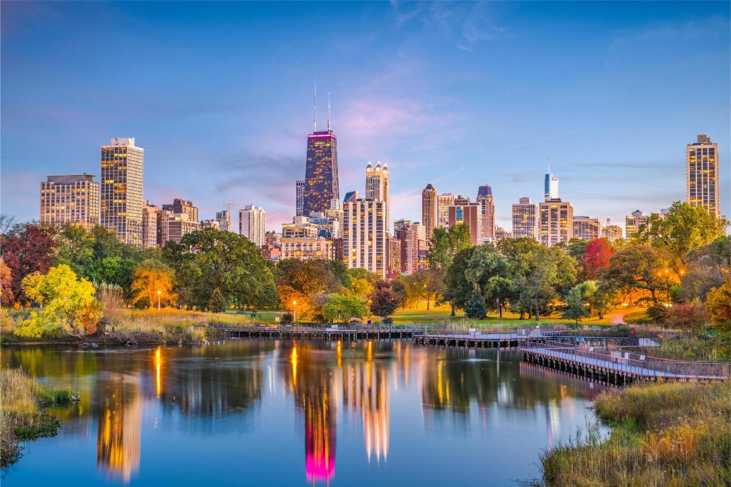 A scenic view of the Chicago skyline from Lincoln Park, featuring high-rise buildings and a body of water in the foreground with a bridge and trees with autumn colors.