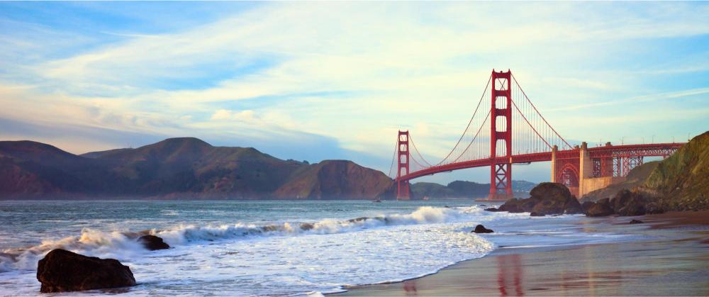 The image shows a view of the Golden Gate Bridge from a beachside perspective, with the bridge spanning the frame and the ocean and hills in the background.