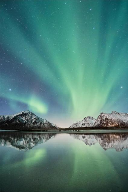 Vibrant green aurora borealis lights up a starry night sky over snowy mountains and their reflection on a calm lake, creating a serene and majestic scene.