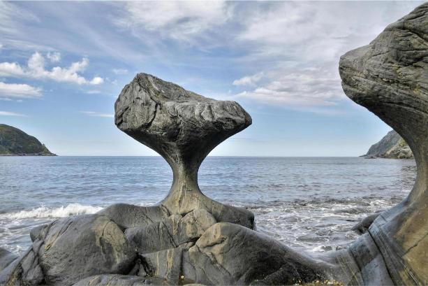 A unique rock formation resembling a pedestal stands by the ocean, eroded at the base. The sky is partly cloudy, with hills visible in the distance.