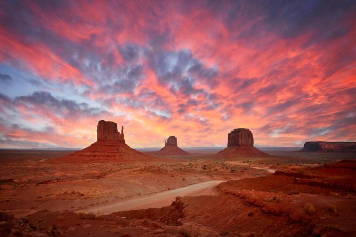 A landscape photograph of the Monument Valley Navajo Tribal Park with sandstone buttes in the background and a dramatic red and orange sky at sunset.