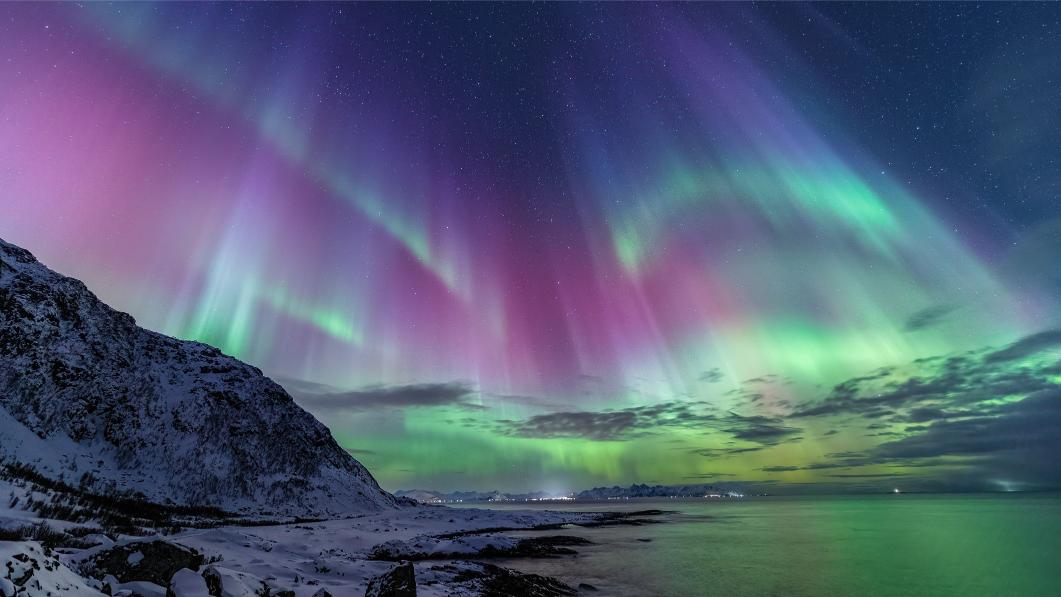 Vibrant northern lights in purple, green, and pink hues illuminate a starry night sky above a snowy mountain and calm sea, creating a serene atmosphere.
