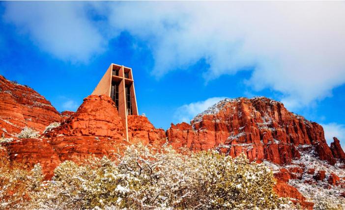 A scenic view of a church-like structure amidst red rock formations in Sedona, Arizona, with a blue sky and some snow on the ground.