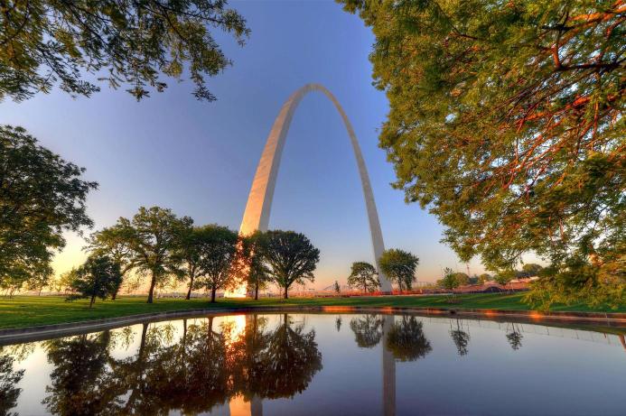 The Gateway Arch in St. Louis, Missouri, reflected in water, with trees and a clear sky in the background.