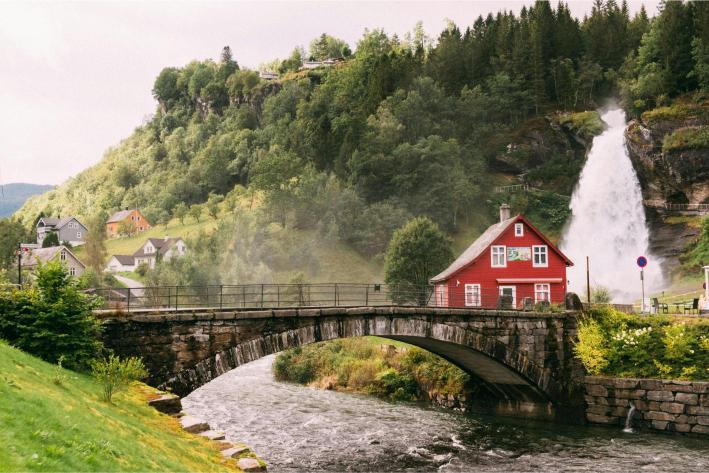A picturesque scene with a red house near a stone bridge over a flowing river. A waterfall cascades in the background, surrounded by lush greenery.