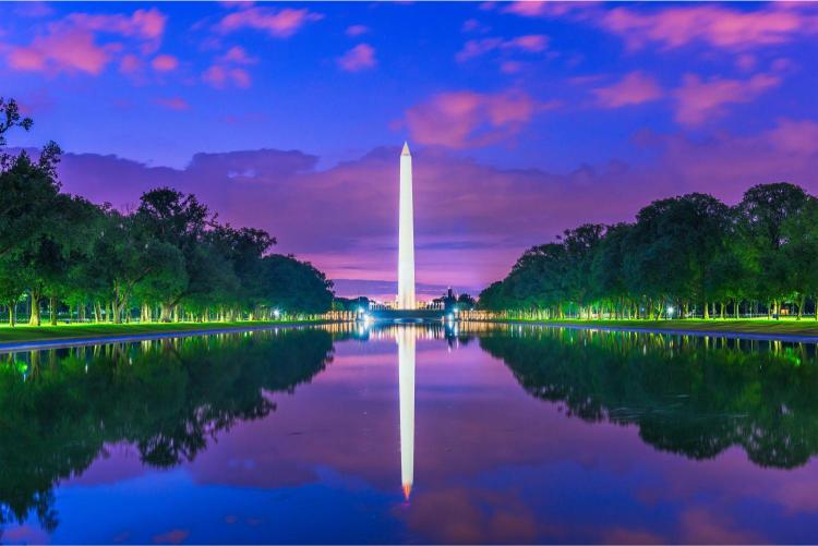 The image shows the Washington Monument in Washington DC, reflecting in a still pool with a colorful sky above.