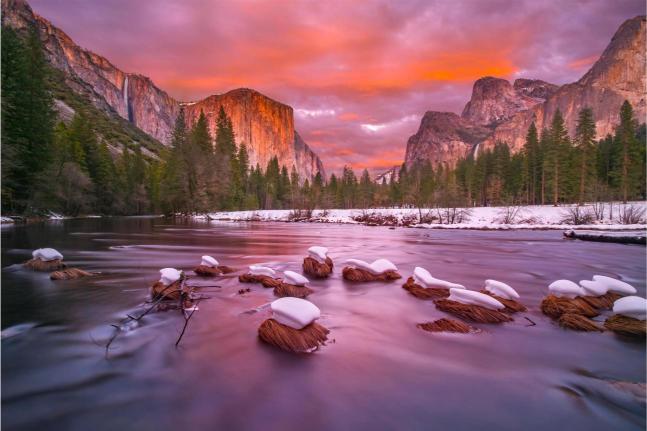 A scenic view of the Yosemite National Park with the Merced River in the foreground and snow-capped mountains in the background, capturing a vibrant sunset.