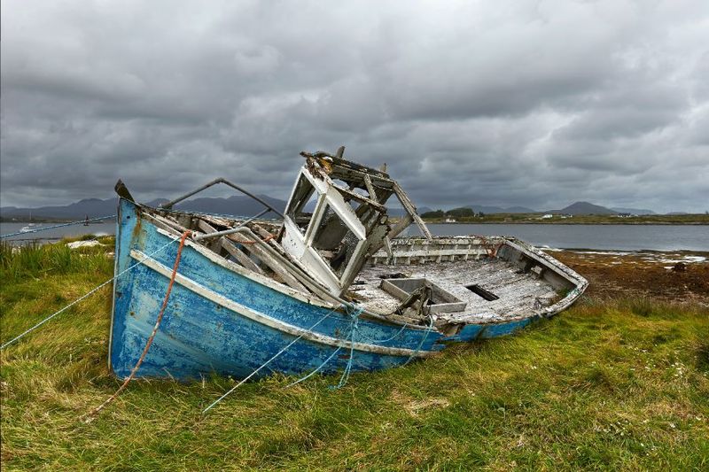 A weathered, abandoned blue boat rests on grassy terrain under cloudy skies, evoking feelings of solitude and decay. Distant mountains line the horizon.