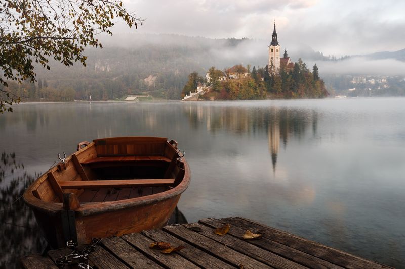 A scenic view of a calm lake with a wooden boat docked at the shore, with a blurred background featuring trees and a castle.