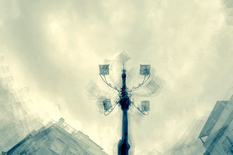 A bird's-eye view of a streetlight set against a cloudy sky, framed by blurred building edges. The image conveys a sense of urban solitude and calm.